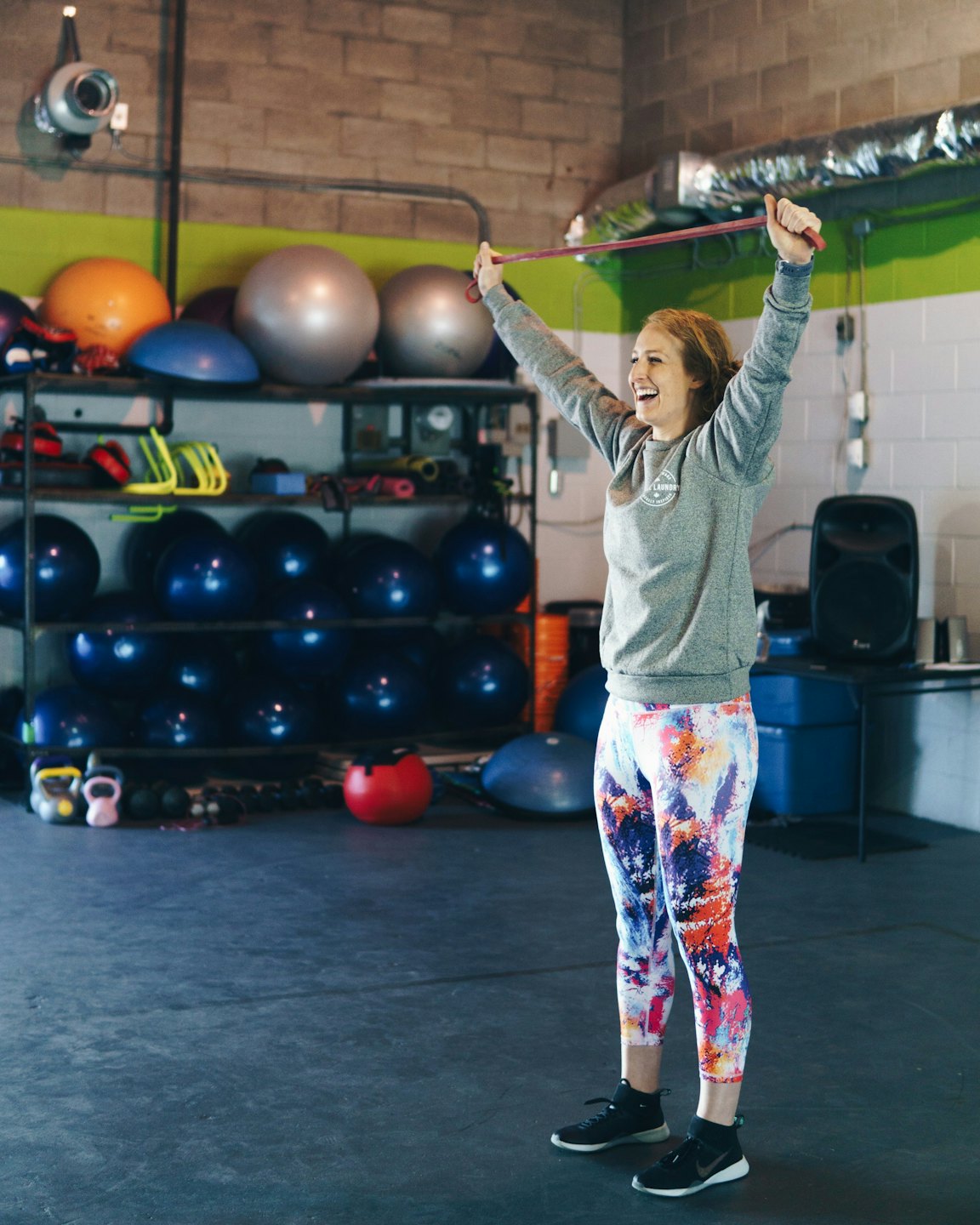 Woman stretching pink resistance band overhead in a gym.