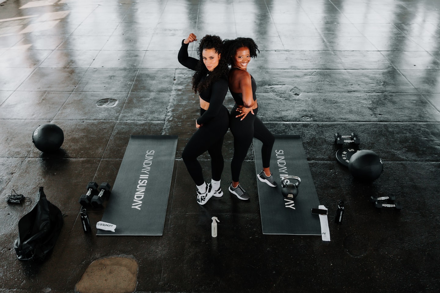Two fit women in black activewear pose back-to-back in a gym.