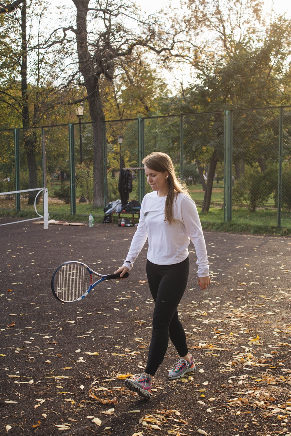 Woman walks across a leaf-covered tennis court with a racket.